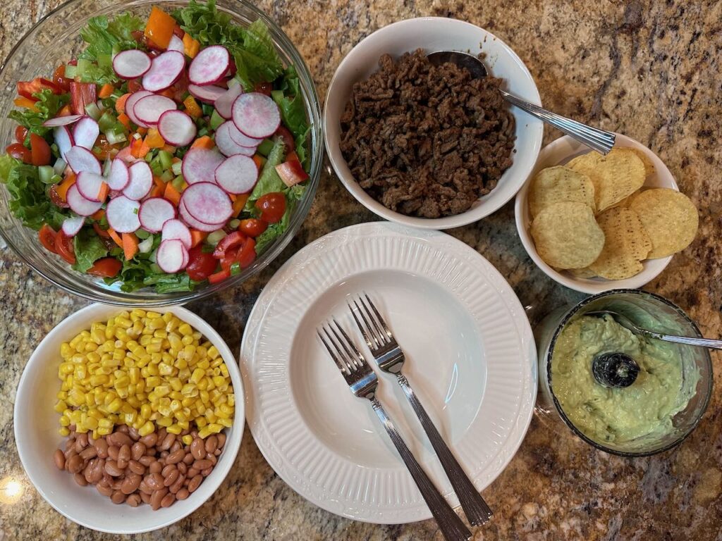 Top view of deconstructed taco salad.  Greens and veggies in one bowl, pinto beans and corn in another, Chips and taco meat (beef) in their own bowls and then the avocado cilantro lime dressing