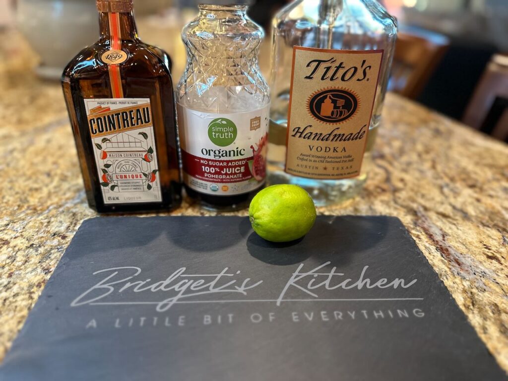 Cointreau, Pom Juice from a bottle, Tito's vodka and a lime.  All in front of a slate slab with the Bridget's Kitchen Logo on it. 