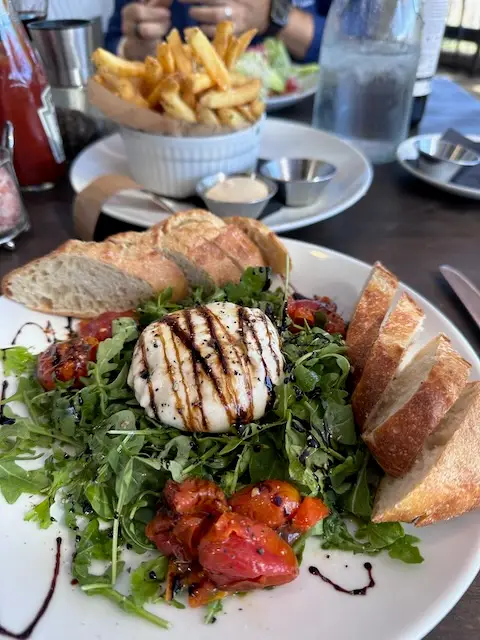 Salad purchased at a restaurant showing baguette bread, buratta, arugula salad and roasted tomatoes.